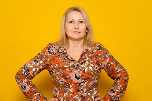 Smiling woman in patterned dress posing isolated on yellow wall background studio portrait. People emotions lifestyle concept. Mock up copy space. Standing with arms akimbo on the waist.