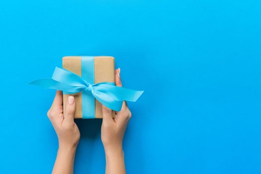 Woman hands give wrapped valentine or other holiday handmade present in paper with blue ribbon. Present box, decoration of gift on blue table, top view with copy space.