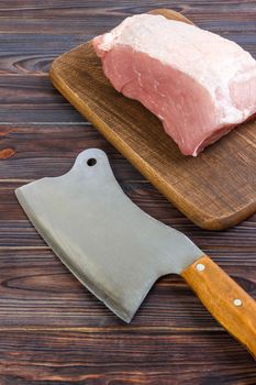 fresh large piece of pork on a wooden cutting board with kitchen ax. Top view on gray concrete background.