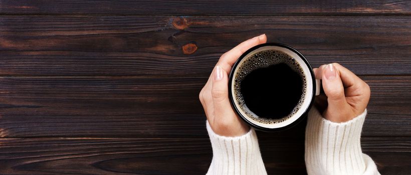 Lonely woman drinking coffee in the morning, top view of female hands holding cup of hot beverage on wooden desk. Banner.