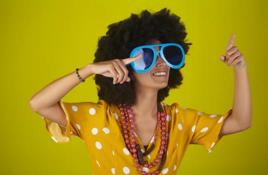 A beautiful and smiling woman with the curly afro hairstyle wearing heart shaped sunglasses looking and indicating direction up to the right with fingers on yellow background