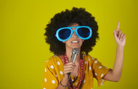 Young beautiful and smiling woman with the curly afro hairstyle and funny sunglasses singing using a microphone on yellow background