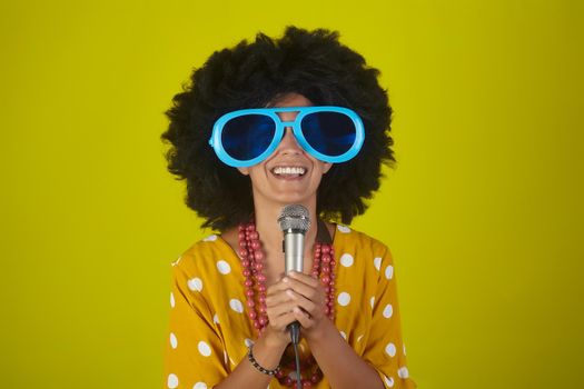 Young beautiful and smiling woman with the curly afro hairstyle and funny sunglasses singing using a microphone on yellow background