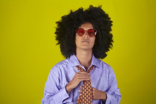 Portrait of a young woman with afro curly hairstyle wearing male clothes straightening a tie on yellow background
