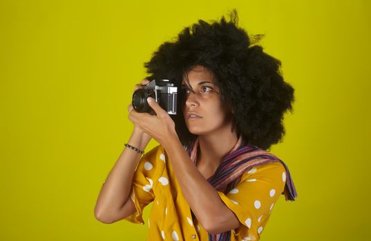 A beautiful woman with curly afro hairstyle on yellow background while taking pictures with a retro films camera