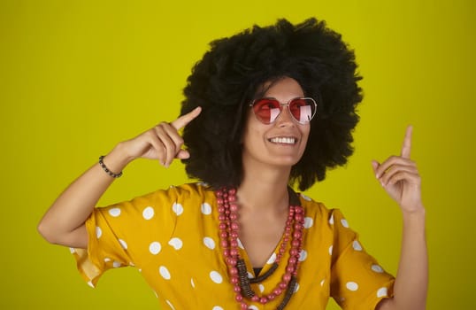 A beautiful and smiling woman with the curly afro hairstyle wearing heart shaped sunglasses looking and indicating direction up to the right with fingers on yellow background