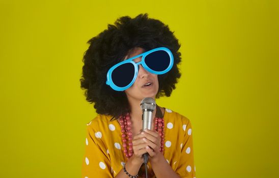 A beautiful woman with the curly afro hairstyle and funny sunglasses singing using a microphone on yellow background