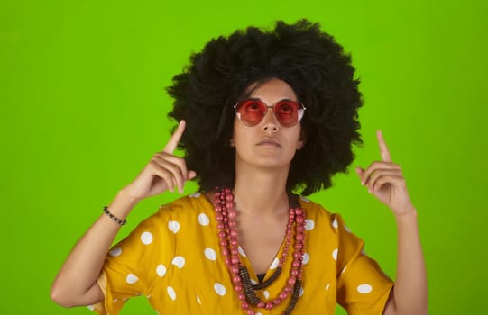 A beautiful woman with the curly afro hairstyle wearing heart shaped sunglasses looking and indicating direction up to the right with fingers on green background