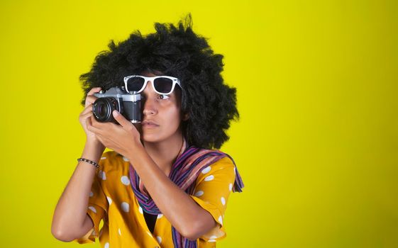 A beautiful woman with curly afro hairstyle on yellow background while taking pictures with a retro films camera