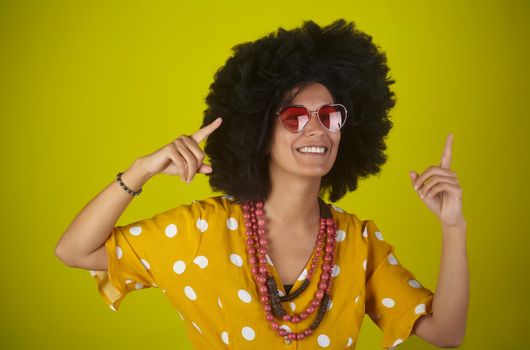 A beautiful and smiling woman with the curly afro hairstyle wearing heart shaped sunglasses looking and indicating direction up to the right with fingers on yellow background