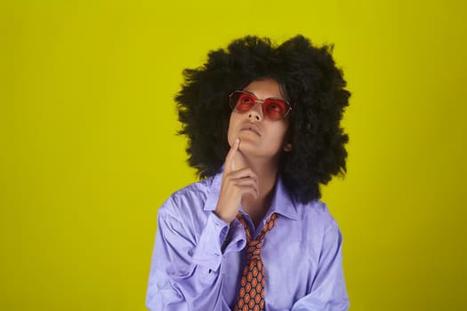 Portrait of a thinking woman with afro curly hairstyle wearing sunglasses, male shirt and necktie on yellow background