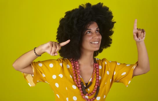 A beautiful and smiling woman with the curly afro hairstyle looking and indicating direction up to the right with fingers on yellow