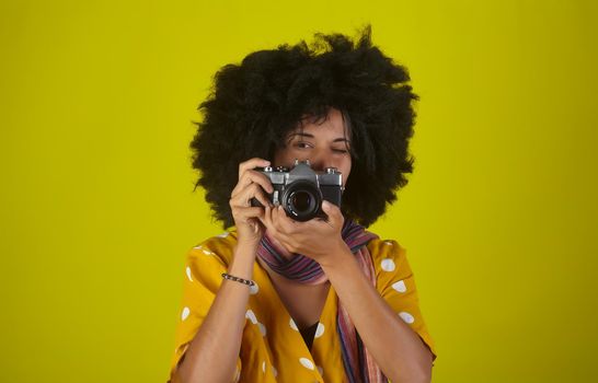 A beautiful woman with curly afro hairstyle on yellow background while taking pictures with a retro camera