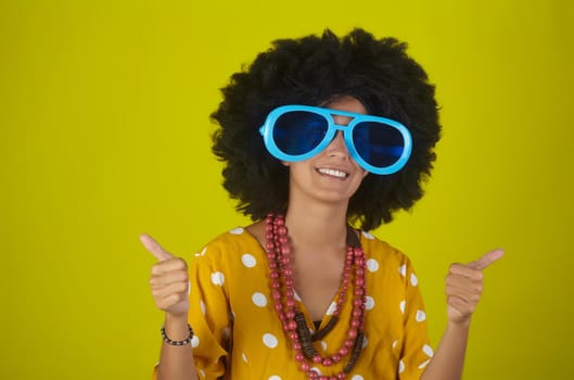 Young beautiful and smiling woman with curly afro hairstyle and funny sunglasses showing thumbs up on yellow background
