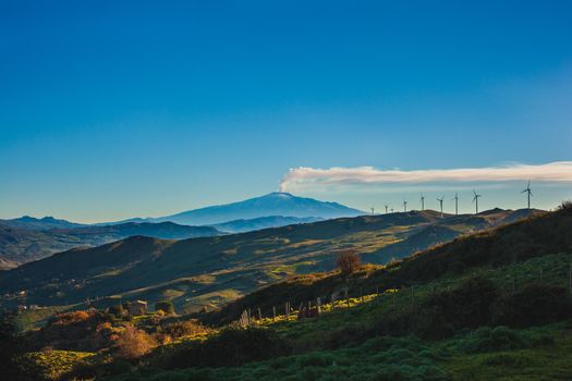 View of windmills and the mount Etna Volcano with smoke. Sicily