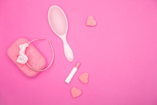 Flat lay of pink female bathroom accessories lying on a pink field. Accessories of every woman including lipstick, hairbrush, sponge, soap, and headband