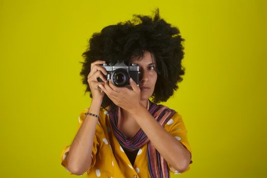 A beautiful woman with curly afro hairstyle on yellow background while taking pictures with a retro camera