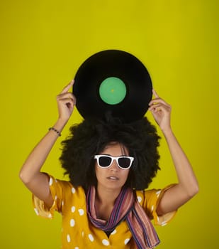 A beautiful woman with sunglasses and afro hairstyle holding on her head a vinyl disk on a yellow background