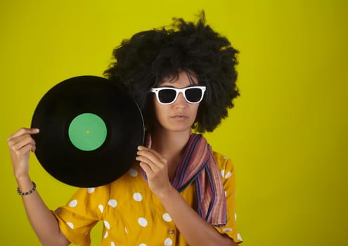 A young woman with sunglasses and afro hairstyle holding a vinyl disk on a yellow background