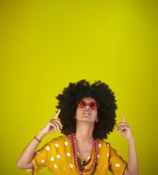 A beautiful woman with the curly afro hairstyle wearing heart shaped sunglasses looking and indicating direction up to the right with fingers on yellow background