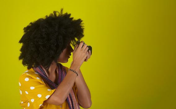 A beautiful woman with curly afro hairstyle on yellow background while taking pictures with a retro films camera