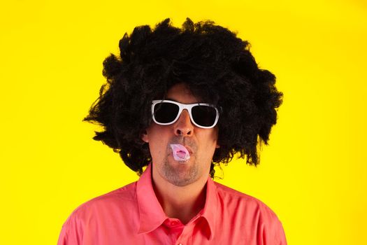 Curly man wearing sunglasses blowing bubble with chewing gum on yellow background