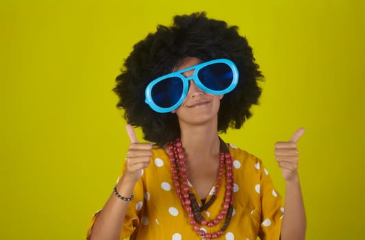 Young beautiful and smiling woman with curly afro hairstyle and funny sunglasses showing thumbs up on yellow background