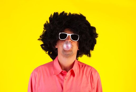 Curly man wearing sunglasses blowing bubble with chewing gum on yellow background