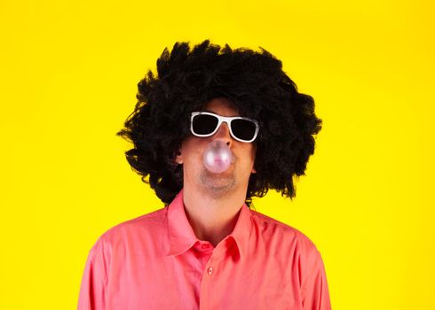 Curly man wearing sunglasses blowing bubble with chewing gum on yellow background