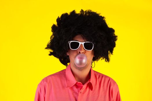 Curly man wearing sunglasses blowing bubble with chewing gum on yellow background