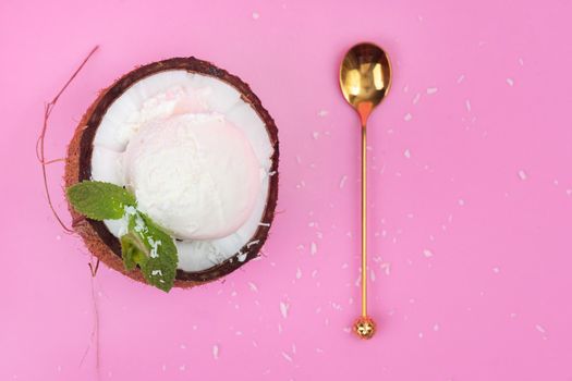 Vanilla ice cream balls in a fresh coconut half decorated with mint leaves with a spoon on a pink background.