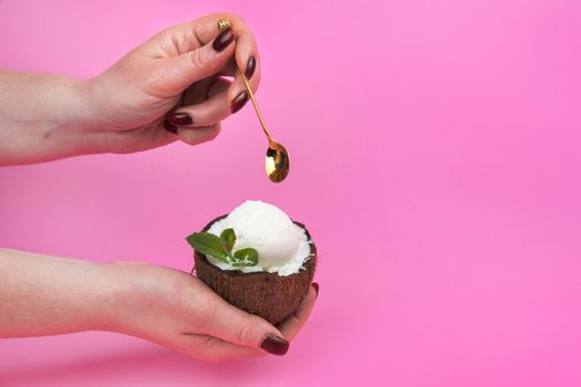 Vanilla ice cream balls in a fresh coconut half, decorated with mint leaves in the hand with a spoon on a pink background.