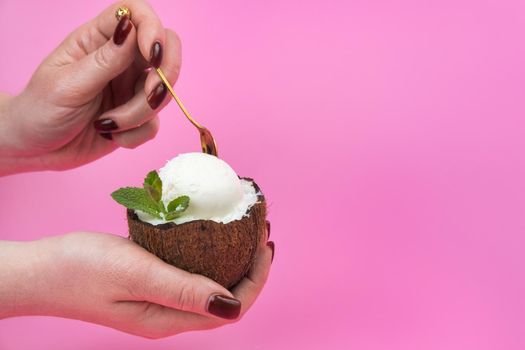 Vanilla ice cream balls in a fresh coconut half, decorated with mint leaves in the hand with a spoon on a pink background.