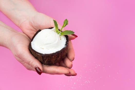 Vanilla ice cream balls in a fresh coconut half, decorated with mint leaves in your hand on a pink background.