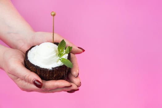 Vanilla ice cream balls in a fresh coconut half, decorated with mint leaves in the hand with a spoon on a pink background.