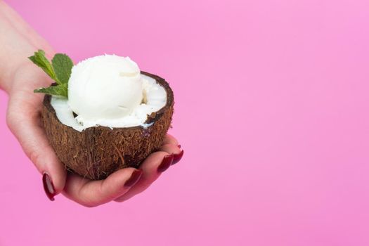 Vanilla ice cream balls in a fresh coconut half, decorated with mint leaves in your hand on a pink background.