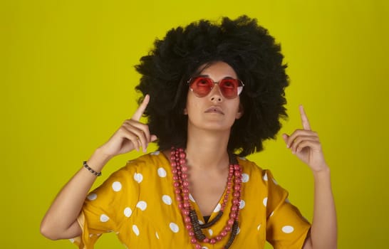 A beautiful woman with the curly afro hairstyle wearing heart shaped sunglasses looking and indicating direction up to the right with fingers on yellow background