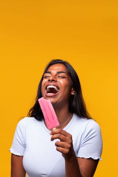 Vertical portrait of young Indian woman laughing eating pink popsicle on yellow background. Asian woman eating ice cream. Studio shot.