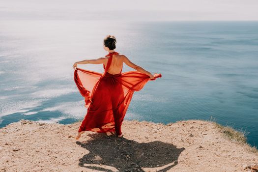 Side view a Young beautiful sensual woman in a red long dress posing on a rock high above the sea during sunrise. Girl on the nature on blue sky background. Fashion photo.