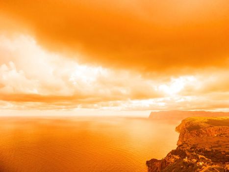 A red burning sunset with the silhouette of a cliff over the sea. Abstract nature summer or spring ocean sea background. Small waves on golden warm water surface with bokeh lights from sun.