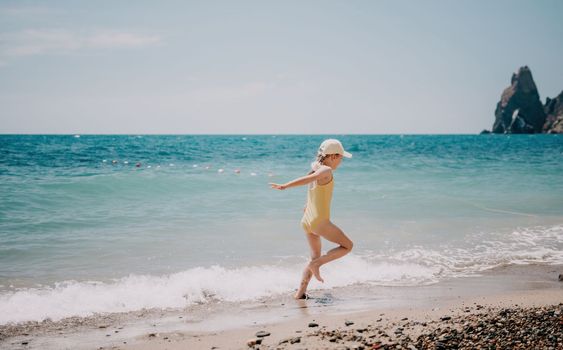 Cute little girl running along the seashore against a clear blue sea and rejoices in the rays of the summer sun. Beautiful girl in yellow swimsuit running and having fun on tropical beach