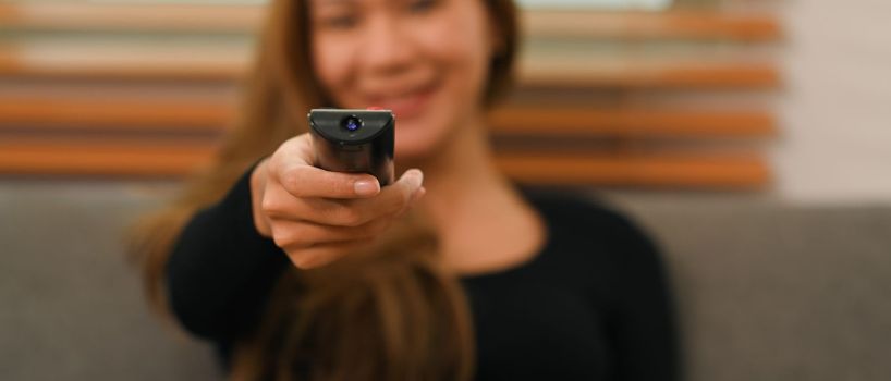 Young woman with remote controller watching TV, relaxing on couch at home. Select focused on hand.