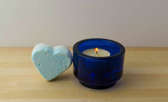 Bath bomb in the shape of a heart and a burning candle on a wooden table.