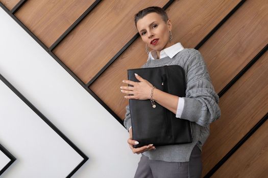 A business woman with a short haircut in a gray sweater with a leather folder for documents in her hands on the background of an office texture wall.