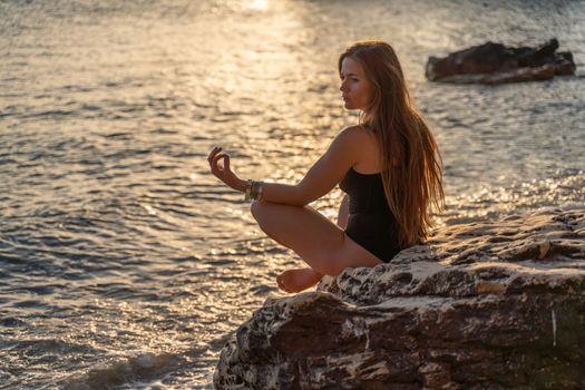 Woman tourist enjoying the sunset over the sea mountain landscape. Sits outdoors on a rock above the sea. She is wearing jeans and a blue hoodie. Healthy lifestyle, harmony and meditation.