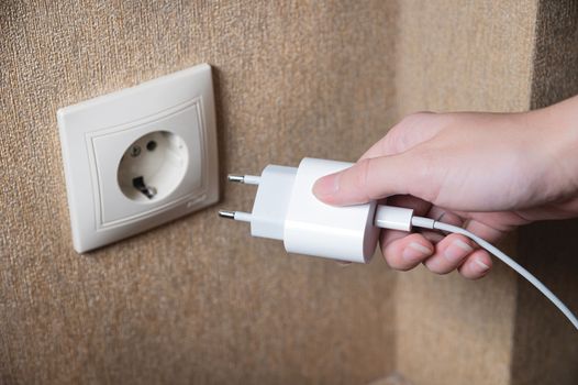 Woman's hand turns on, turns off the charger in the electrical socket on the wall in the house, close-up.