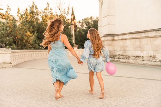 Daughter mother run holding hands. In blue dresses with flowing long hair against the backdrop of a sunset and a white building