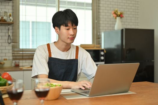 Smiling asian man looking at laptop screen, working online while sitting at wooden table in kitchen.