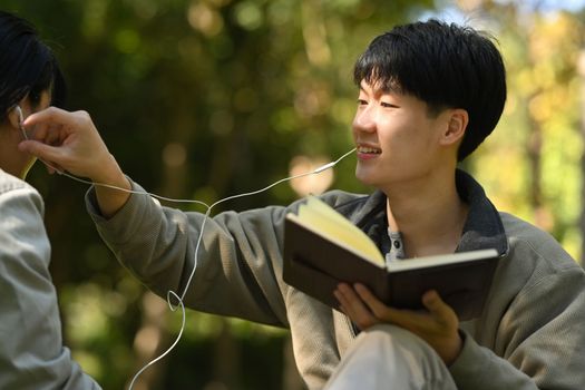 Image of young couple reading a book and listening to music in earphones while relaxing at public park together.