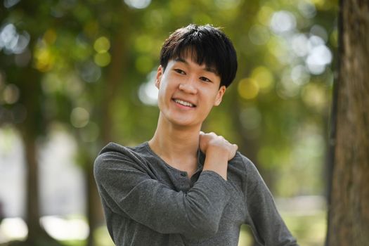 Pleasant asian man in casual clothes relaxing, stretching arms while standing in the park outdoors.